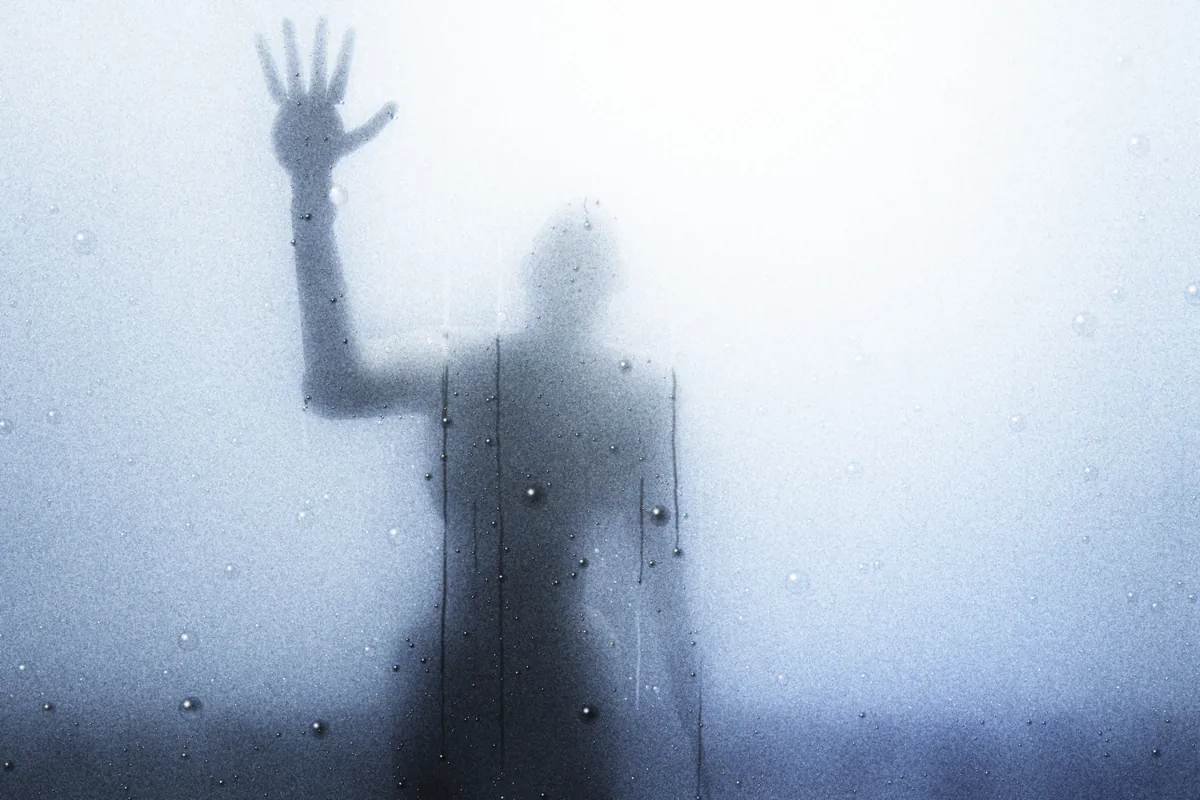 Une silhouette floue derrière une vitre embuée, la main posée à plat contre le verre ; gouttes et coulures de condensation dans une lumière froide.