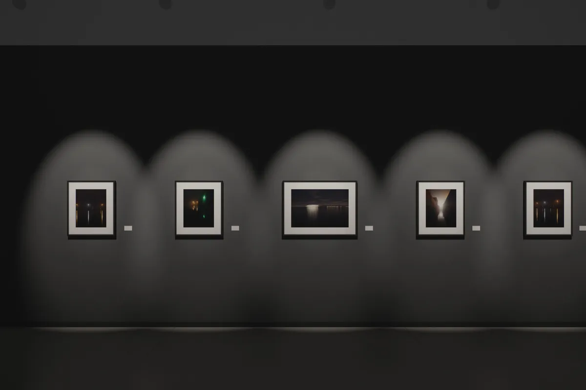 Vue d’exposition : cinq tirages de la série « Nocturnes portuaires » encadrés de noir sur un mur gris anthracite, chacun éclairé par un projecteur, au-dessus d’un sol en béton ciré.