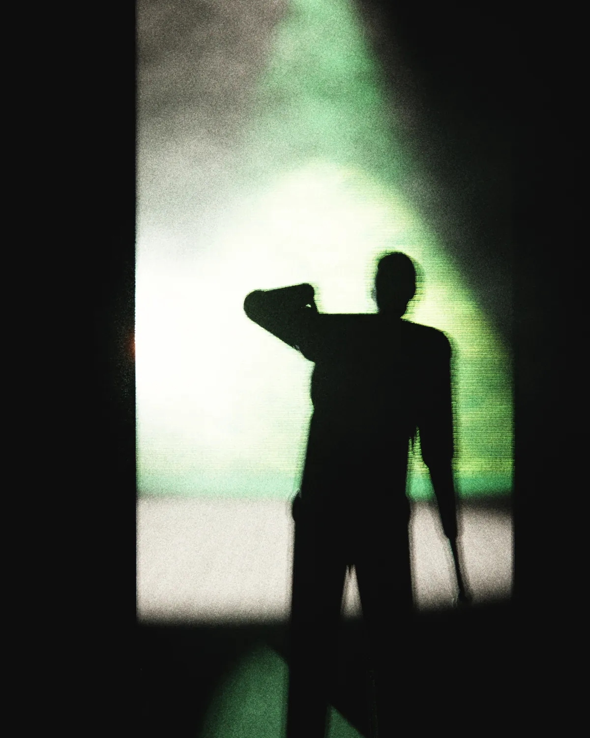 Silhouette d’homme en contre-jour dans une coulisse, de dos, une main sur la nuque et un micro dans l’autre, face au plateau éclairé.