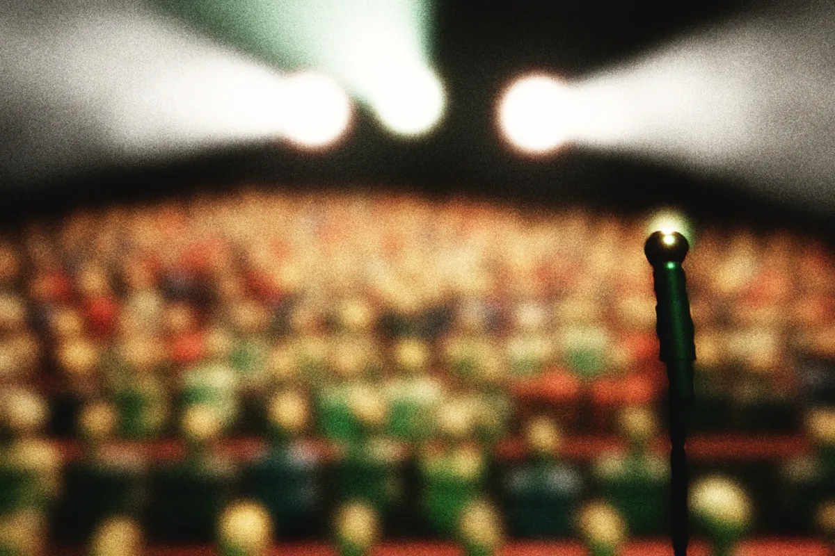 La salle comble vue depuis la scène : rangées de spectateurs floues sous la lumière de salle, projecteurs de face éblouissants au fond et pied de micro au premier plan.
