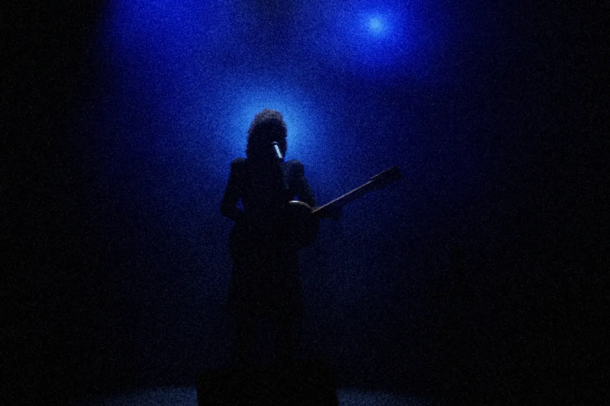 Romy Calvel seule au micro avec sa guitare, en silhouette : un projecteur bleu caché derrière sa tête dessine un halo dans la brume de scène.